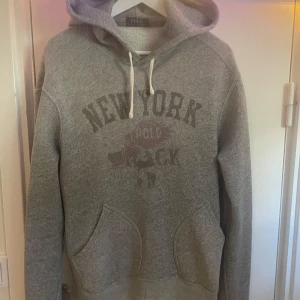 Grå hoodie från Polo Ralph Lauren - Säljer en grå hoodie från Polo Ralph Lauren i storlek M. Tröjan har tryck med texten 'New York' på bröstet, huva med snörning och en stor magficka. Perfekt för chill dagar och har en klassisk streetstil. Materialet är mjukt och känns som bomull.