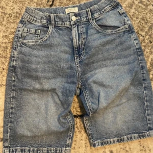 Blå jeansshorts från Lindex, 164 - Snygga blå jeansshorts från Lindex i klassisk modell med fem fickor och knappstängning. Shortsens längd går strax ovanför knäet och de har coola bakfickor med lock och knapp. Perfekta för dig som gillar avslappnad streetstil.