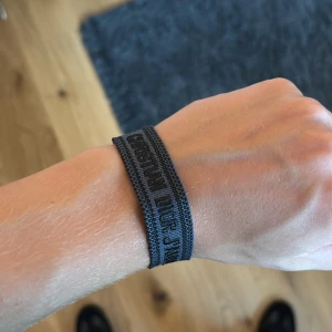 Christian Dior J'Adior armband i svart - Snyggt svart armband från Christian Dior med texten 'J'Adior' broderad i mörkgrått. Armbandet är gjort i textil och har fransar i ändarna för en cool touch. Perfekt accessoar för att lyfta din outfit med lite lyxig vibe.