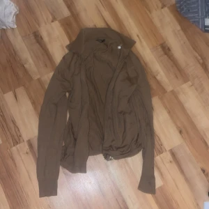 Brun kofta från J.Lindeberg med dragkedja - Snygg brun kofta från J.Lindeberg med dragkedja framtill och hög krage. Materialet är mjukt och stickat, perfekt för lager på lager under höst och vinter. Långärmad modell med clean look som funkar till jeans eller chinos.