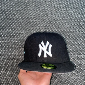 Svart New Era Yankees keps - Snygg svart New Era 59FIFTY keps med New York Yankees-logga i vitt framtill. Broderad frihetsgudinna och texten 'Greetings from New York' i vitt och blått på sidan. MLB-logga bak och New Era-logga på sidan. Klassisk flat peak och stilren look.