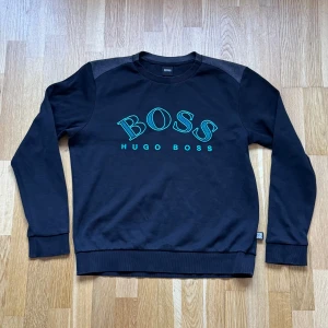 Hugo Boss Svart Crewneck - Hugo Boss Crewneck med broderat tryck fram i färgen svart. OBS! En tråd har gått upp lite i nacken men inget som syns när den är på! Storlek: L, Armhåla till Armhåla: 56 cm, Längd: 62 cm, Axel till Armöppning: 60 cm.  Material: 83% Cotton, 11% Cotton (Recycled), 6% Polyamid. 