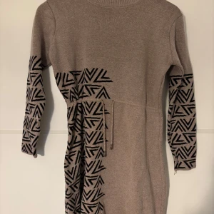 Beige stickad klänning med mönster - Snygg beige stickad klänning med svarta grafiska mönster på ärmar och ena sidan. Klänningen har lång ärm och ett knytband i midjan för extra stilpoäng. Perfekt för dig som gillar unika detaljer och mjuka material.