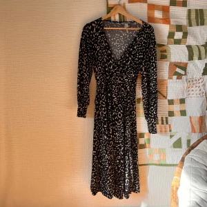Svart leopardmönstrad klänning - Snygg svart långklänning från H&M med leopardmönster i beige och grå toner. Klänningen har omlott-design med v-ringning, långa ärmar och knyt i midjan. 