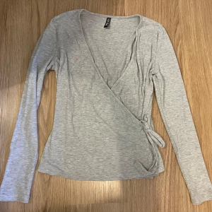 Ljusgrå omlottopp från Pieces - Supersnygg ljusgrå omlottopp från Pieces med långa ärmar och knyt i sidan. Toppens ribbade material ger en skön känsla och den v-ringade designen gör den extra fin. Står att det är st xs men passar mer M/L definitivt.