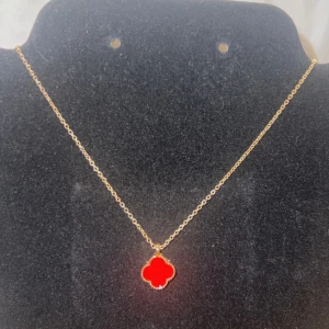 Guldhalsband med röd fyrklöverberlock - Snyggt halsband i guld med en röd fyrklöverformad berlock som hänger i en tunn kedja. Perfekt för dig som gillar färgstarka detaljer och vill sticka ut med något unikt. Berlocken har mjuka kanter och ger en cool och modern vibe.