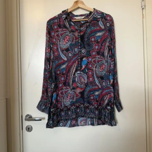 Mönstrad långärmad skjorta från Indiska - Färgstark långärmad skjorta från odd Molly med paisley- och blommönster i blått, rött och vitt. Skjortan har knappar framtill och en lös, flowy passform. Perfekt för dig som gillar boho-vibe och vill sticka ut med coola mönster. Storlek 2 som enligt deras storleksguide motsvarar en storlek M.  Mycket fint skick, jag hittar inga synliga anmärkningar eller defekter.
