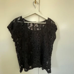Svart blommig spetsblus - Säljer en svart blus i genombruten spets med blommigt mönster. Toppen har rund halsringning/off sholder och korta ärmar. Perfekt att styla lager på lager eller med ett linne under för en cool look. Säljer för att jag ej använder längre, jag har ofta xs/s i toppar💗