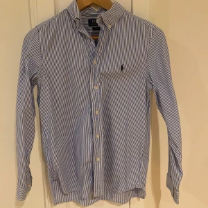 Randig skjorta från Polo Ralph Lauren - Skjortan är i ny skick. Snygg blå- och vitrandig skjorta från Polo Ralph Lauren i slim fit-modell. Skjortan har klassisk krage, knäppning framtill och broderad logga på bröstet. Tillverkad i mjuk bomull och har långa ärmar med manschettknappar.