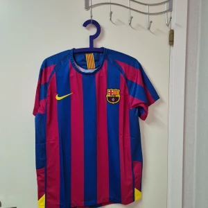 Barcelona Ronaldinho 10 fotbollströja - Säljer en klassisk FC Barcelona matchtröja från Nike med Ronaldinho och nummer 10 på ryggen. Tröjan har breda blå och röda ränder, gula detaljer och klubbmärke på bröstet. Kortärmad och tillverkad i lätt polyester som andas. Perfekt för dig som älskar fotboll och legender.