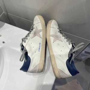 Golden Goose Superstar vita sneakers - Golden Goose Superstar sneakers i vitt skinn med grå mockastjärna på sidan, blå hälkappa och grå snörning. Klassisk rund tå och platt sula. Snygg kontrast mellan material och färger, perfekt för en trendig streetstyle-look.