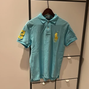 Turkos Polo Ralph Lauren piké, slim fit - Snygg turkos pikétröja från Polo Ralph Lauren i slim fit. Klassisk krage, korta ärmar och stora gula broderier – logga på bröstet och siffran 3 på ärmen. Tillverkad i bomull, perfekt för en fräsch och sportig look.