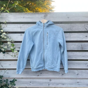 Ljusblå hoodie från Sail Racing - Ljusblå hoodie från Sail Racing med dragkedja hela vägen, huva och en bröstficka med dragkedja. Broderad logga på bröstet och mjukt fleecematerial på insidan. Perfekt för kyliga dagar och snygg till jeans eller joggers.