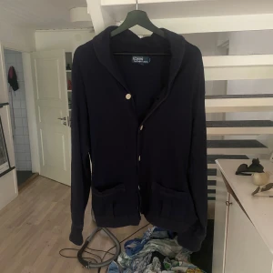 Mörkblå kofta från Polo Ralph Lauren - Säljer en mörkblå kofta från Polo Ralph Lauren i storlek L. Koftan har huva, två stora fickor framtill och stängs med knappar. Tillverkad i mjuk bomull, perfekt för chill dagar. Klassisk och enkel design som funkar till det mesta.