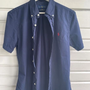 Mörkblå kortärmad skjorta Ralph Lauren - Mörkblå kortärmad skjorta från Ralph Lauren i slim fit-modell. Skjortan är tillverkad i tunn bomull och har klassisk krage, vita knappar och röd broderad logga på bröstet. Perfekt för dig som gillar stilren och tidlös design.