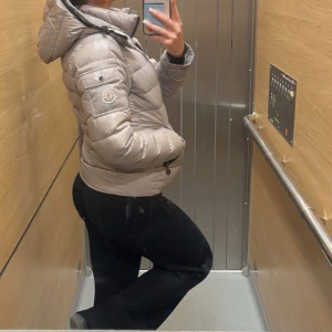 Grå dunjacka från Moncler - Säljer en snygg grå dunjacka från Moncler med huva och dragkedja. Jackan är quiltad och har en klassisk Moncler-logga på ärmen. Perfekt för kyliga dagar och har en modern, lite kortare passform.