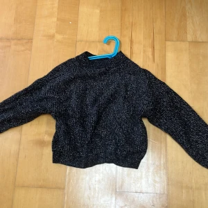 Svart glittrig stickad tröja - Svart stickad tröja med silvriga glittertrådar som ger en snygg shimmer-effekt. Tröjan har lång ärm, rak passform och lite högre hals. Perfekt för dig som vill ha något extra till din outfit. Mjuk och skön att bära.