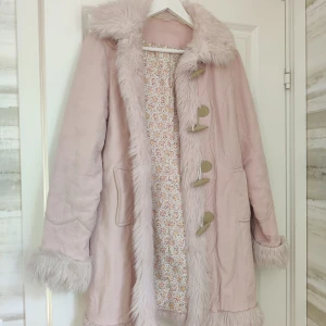 Ljusrosa penny lane kappa med broderi - Superfin vintage ljusrosa penny lane kappa med fuskpälsdetaljer på krage, ärmslut och längs öppningen. Har ett broderat+tryckt mönster nertill, tyget är väldigt likt manchester tyg. Tunt foder, definitivt en vår och höstjacka. Säljs as is, jag köpte den asdyrt vintage och har fått bort parfymdoften men inte haft tillfälle att använda den själv. Blommigt foder. 