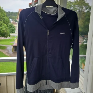 Mörkblå hoodie från BOSS med dragkedja - Snygg mörkblå hoodie från BOSS med grå detaljer på krage, muddar och nederkant. Tröjan har hel dragkedja, fickor framtill och diskret BOSS-logga på bröstet. Perfekt för chill dagar och enkel att matcha med jeans eller joggers. Jag ryckte lappen o va tvungen att sy ihop men tröjan är i princip oanvänd