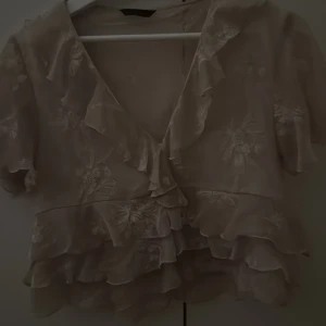 Rosa blommig blus med volanger - Supersöt rosa blus med broderade blommor och volangdetaljer. Den har v-ringning, korta ärmar och flera lager volanger nertill. Tunn och luftig, perfekt för vår och sommar. Ger en romantisk och drömmig vibe till din outfit.