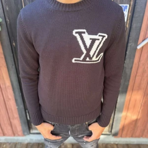 Mörkgrå stickad Louis Vuitton tröja - Snygg mörkgrå stickad tröja från Louis Vuitton med stort vitt LV-logo på bröstet. Rund hals och långa ärmar med ribbade muddar. Perfekt för dig som gillar exklusiva märken och vill ha en clean look med tydlig branding.