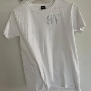 Vit t-shirt med strass från Bvaldi - Vit t-shirt från Bvaldi i bomull, med korta ärmar och rund hals. Framsidan har ett stort BV i strass och baksidan har texten 'Need money for Porsche' i strass. Snygg och edgy stil, perfekt för dig som gillar statement-plagg. Ny skick utan pris lapp aldrig använd 