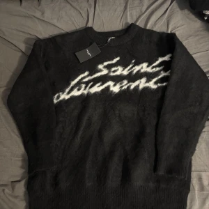 Svart Saint Laurent stickad tröja - Exklusiv svart stickad tröja från Saint Laurent med vit logotyp i grafisk stil över bröstet. Tröjan har lång ärm, rund halsringning och är tillverkad i ett fluffigt, mjukt material som känns lyxigt. Perfekt för dig som vill ha en statement piece i garderoben.