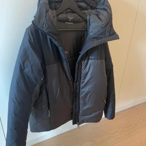 Svart dunjacka från Houdini med huva - Säljer en svart dunjacka från Houdini med stor, mysig huva och dragkedja framtill. Jackan har en clean look, två sidofickor med dragkedja och är perfekt för kalla dagar. Materialet är lätt och smidigt, vilket gör den enkel att bära och styla till olika outfits.