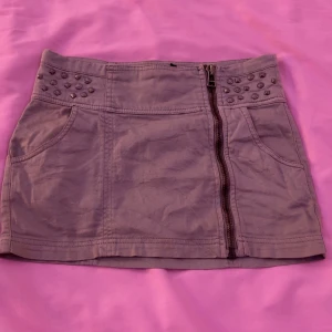 Grå minikjol med nitar från TRF - Säljer en grå minikjol från TRF med dekorativa nitar vid linningen och dragkedja framtill. Kjolen har två fickor fram och är sydd i ett mjukt bomullstyg. Perfekt för dig som gillar detaljer och vill sticka ut lite extra.