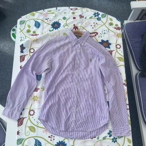 Lila och vit randig skjorta från Polo Ralph Lauren - Säljer en slim fit skjorta från Polo Ralph Lauren i lila och vitt med klassiska ränder. Skjortan har lång ärm, knappar framtill och den ikoniska gröna broderade loggan på bröstet. Perfekt för dig som gillar stilrena plagg med snygg passform.