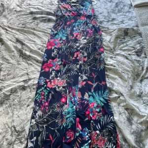 Blommig långklänning med omlott - Snygg långklänning med tropiskt blommönster i rosa, blått och grönt på marinblå botten. Klänningen har omlottliv och smala axelband, perfekt för sommarens alla tillfällen. Är osäker på storleken men mellan 40-50 skulle jag gissa på