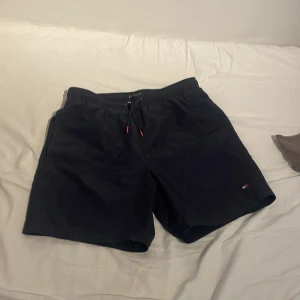 Marinblå badshorts från Tommy Hilfiger - Säljer ett par marinblåa Tommy Hilfinger bad shorts då de är försmå för mig. Dem har två sido fickor samt en bak ficka (se bild 2). Har använts en del men fortfarande i fint skick. Storlek 12-14 år. Perfekta inför sommaren, tveka inte att höra av dig vid frågor eller mer bilder :)