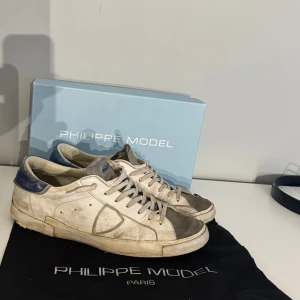 Vita sneakers från Philippe Model med blå detaljer - Säljer ett par vita Philippe Model sneakers med blå detaljer vid hälen och grå mocka vid tån. Skorna har snörning och en platt sula med märkets logga undertill. Perfekta för dig som gillar stilrena och klassiska sneakers. Finns digitalt kvitto och skorna är lite slitna vid sulan men fullt användbara. Box och dustbag finns