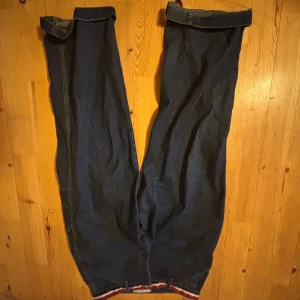 Blå jeansbyxor från Tommy Hilfiger - Säljer ett par klassiska blå jeansbyxor från Tommy Hilfiger med raka ben och uppvikta benslut. Byxorna har bakfickor och synliga sömmar i kontrastfärg. lite heelbite längst ner pågrund av uppvikningen