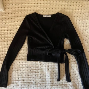 Svart omlottopp med knyt från NA-KD - Säljer en svart ribbad omlottopp från NA-KD i storlek XS. Toppen har långa ärmar, v-ringning och knyt i midjan. Tröjan har knappt använts och är i mycket bra skick.