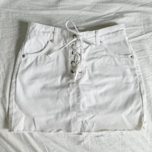 Vit jeanskjol med snörning från Topshop - Säljer en vit jeanskjol från Topshop med snörning framtill och dragkedja bak. Kjolen har rå kant nertill och klassiska fickor fram. Påminner om de populära vita jeansen ifrån Zara