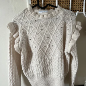Vit stickad tröja med volang  - Superfin vit stickad tröja från Primark med volangdetaljer på axlarna och diskreta pärlor framtill. Tröjan har ett snyggt diamantmönster och ribbade muddar vid ärmslut och nederkant. Perfekt för kyliga dagar! Köptes för ca 1,5 år sedan och har använts en gång efter det!