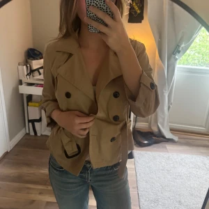 Beige trenchcoat med bälte - Snygg beige trenchcoat med dubbelknäppning och bälte i midjan. Bältet går att ta av, väldigt bra skick, någon liten fläck jag ej testat ta bort, men som ej syns 
