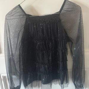 Svart glittrig blus  - Säljer en jättefin svart, glittrig blus! Den har volanger på framsidan och genomskinliga armar. 💕💕