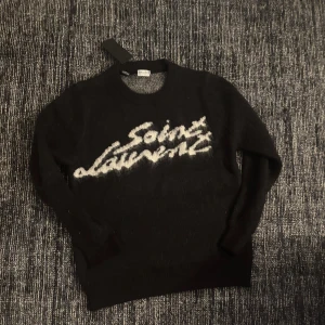 Svart stickad tröja från Saint Laurent - Snygg svart stickad tröja från Saint Laurent med vit text på framsidan. Tröjan har långa ärmar och ribbade muddar vid ärmslut och nederkant. 