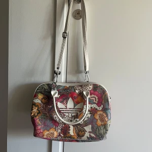 Blommig axelväska från Adidas - Säljer en färgglad axelväska från Adidas med stort vitt logotryck framtill. Väskan har blommigt mönster i rosa, gult, grönt och orange samt vita handtag och axelrem. Perfekt för dig som vill sticka ut med en unik accessoar.