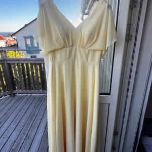 Ljusgul långklänning med v-ringning - Elegant ljusgul långklänning med v-ringad hals och vida, korta ärmar. Klänningen har en mjukt fallande kjol och markerad midja. Perfekt för sommarens speciella tillfällen eller till en studentbal.