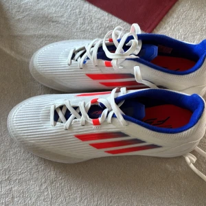 Vita Adidas F50 fotbollsskor med röda och blå detaljer - Säljer ett par vita Adidas fotbollsskor med röda ränder på sidorna och blå insida. Skorna har snörning och en platt sula med mönster för bra grepp. Perfekta för dig som vill sticka ut på planen!