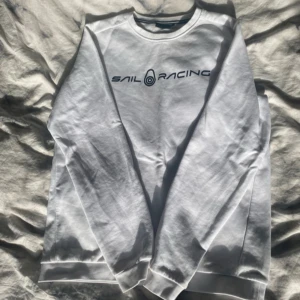 Vit sweatshirt från Sail Racing - Vit sweatshirt från Sail Racing med svart logotyptryck på bröstet. Tröjan har rund halsringning och långa ärmar. Tillverkad i mjuk bomull med ribbade muddar vid ärmslut och nederkant. Perfekt för en avslappnad stil.