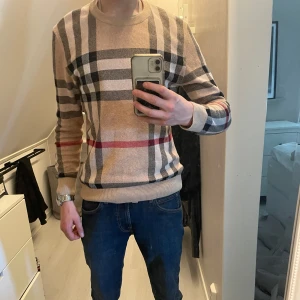 Beige rutig tröja från Burberry - Säljer en klassisk beige tröja från Burberry med det ikoniska rutmönstret i svart, vitt och rött. Tröjan har rund halsringning och långa ärmar. Perfekt för dig som gillar stilrena och tidlösa plagg.