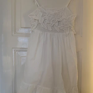 Vit klänning med virkad överdel från Next - Söt vit klänning från Next med smala axelband och virkad överdel med blommönster. Klänningen är luftig och har volang nertill. Perfekt för varma dagar och somriga tillfällen.