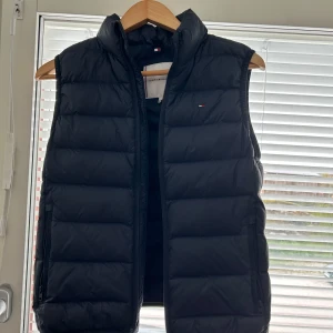 Marinblå dunväst från Tommy Hilfiger - Säljer en marinblå dunväst från Tommy Hilfiger med diskret logga på bröstet. Västen har dragkedja framtill och är quiltad för extra värme. Perfekt att bära över en tröja under kyliga dagar.