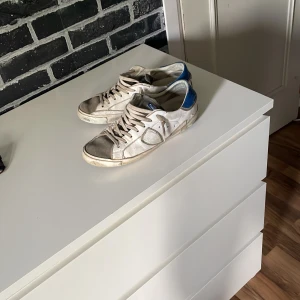 Vita sneakers med blå detalj - Säljer ett par vita sneakers från Philip model med blå häl och grå detaljer. Skorna har snörning och klassisk rund tå. Perfekta för dig som gillar en avslappnad och trendig stil. Dem är lite använda men om man gör rent dem så ser dem nog riktigt bra ut👍 är storlek 41-42👍
