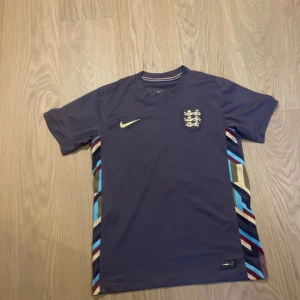 England bortatröja Bellingham 10 Nike - Säljer en England bortatröja från Nike med Bellingham 10 på ryggen. Tröjan är mörklila med färgglada ränder längs sidorna och har det klassiska England-emblemet på bröstet. Perfekt för dig som vill sticka ut på läktaren eller planen.
