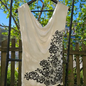 Vit topp med svart blommigt mönster - Säljer en vit topp med snygg svart blommig detalj längs ena sidan. Toppen har en djup v-ringning och är ärmlös, perfekt för varma dagar. Materialet känns lätt och svalt.